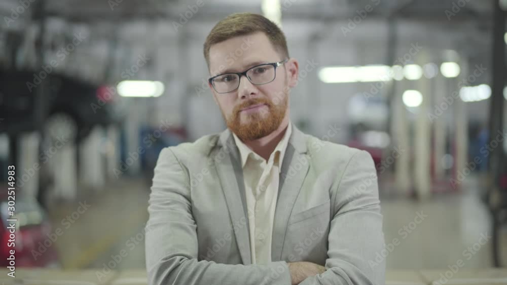 Portrait of confident Caucasian man with red beard crossing arms and looking at camera. Successful car dealer in elegant eyeglasses satisfied with his work. Car dealership, car business. Portrait of confident Caucasian man with red beard crossing arms and looking at camera. Successful car dealer in elegant eyeglasses satisfied with his work. Car dealership, car business.