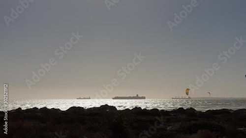 Kites of Cape Town kitesurfers flying behind beach vegetation. Boat silhouette on horizon. Robben Island  visible on right.  Sun glistening on silver ocean waves. Sunlight on mirror sea. Vacation