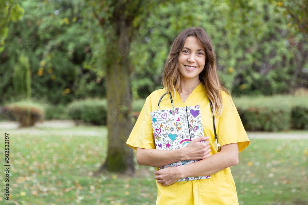 Mujer enfermera joven y sonriente con uniforme amarillo sujetando una carpeta con corazones en entorno natural Mujer enfermera joven y sonriente con uniforme amarillo sujetando una carpeta con corazones en entorno natural