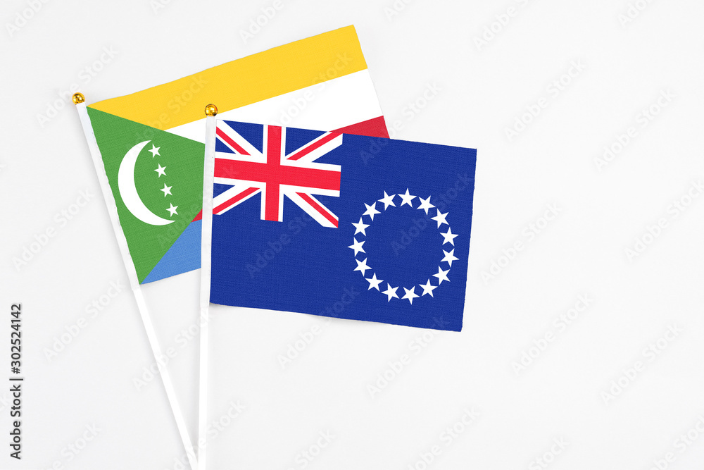 Fototapeta premium Cook Islands and Comoros stick flags on white background. High quality fabric, miniature national flag. Peaceful global concept.White floor for copy space.