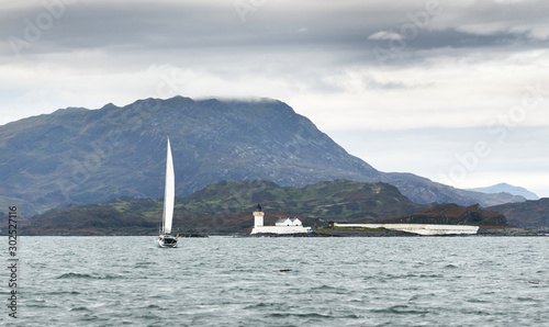 Panoramic view of the cliffs, mountains and valleys of the islands of Inner Hebrides on a cloudy day. Scotland, UK. White sloop  rigged yacht and old loghthouse close-up photo