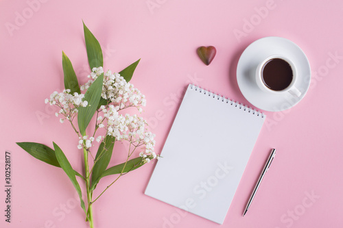 Workspace with espresso cup, paper notebook, pen and gypsophila flowers on pink background