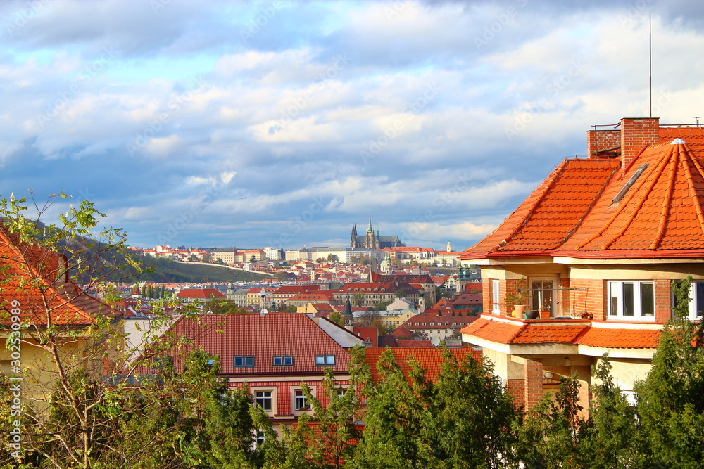 Houses with orange roofs behind green trees against the background of the old city of Prague, the Cathedral in the distance against the blue sky with purple and white clouds.