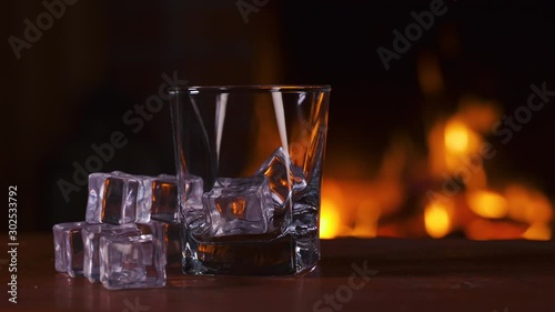 Empty glass with ice stands on a wooden table against the background of a fireplace with flame. Alcoholic drink concept. Fire background. Drink alone. Macro. Close up.