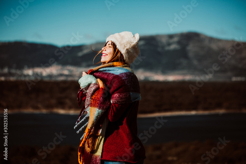 .Pretty young woman enjoying a sunny winter day outdoors. Surrounded by nature, relaxed and carefree. Covering from the cold with a burgundy scarf and wearing a fur hat. Lifestyle