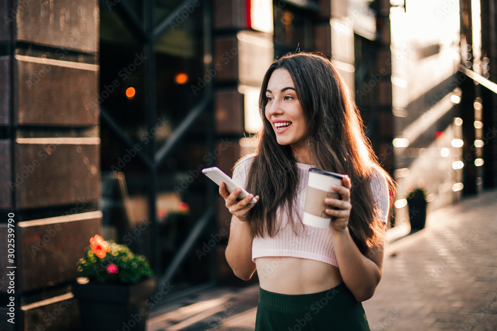 Fototapeta premium Smiling woman using phone and drink coffee on the street in summer day.