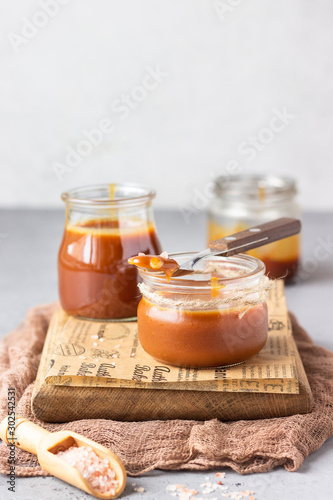 Homemade salted caramel sauce in glass jars on wooden cutting board. Selective focus.