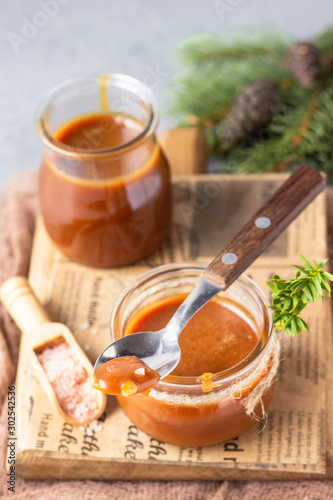 Spoon with tasty caramel sauce over glass jar as a gift for Christmas or New year with fir branches and cones. Selective focus.
