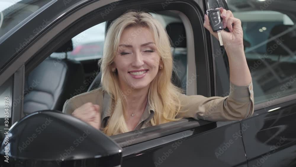 Satisfied Caucasian blond woman with brown eyes bragging car keys while sitting in the salon. Successful businesslady buying automobile in showroom. Car dealership, car business. Satisfied Caucasian blond woman with brown eyes bragging car keys while sitting in the salon. Successful businesslady buying automobile in showroom. Car dealership, car business.