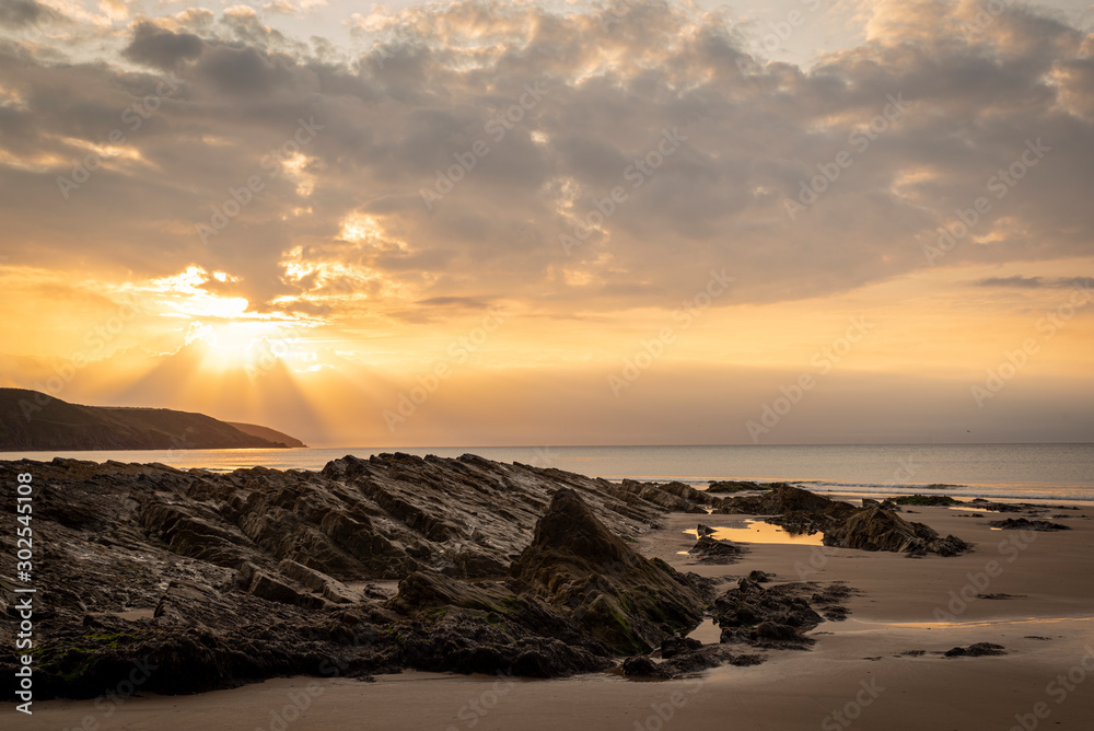 sunrise with golden light beams, rocks in foreground with sand,  with yellow sky and clouds