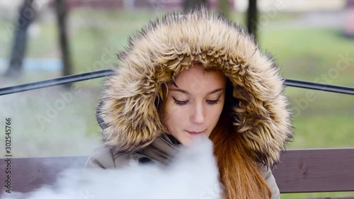 Vaping teenager. Young pretty white caucasian teenage girl in green jacket with hood smoking an electronic cigarette on the bench on the street in the autumn day. Bad habit. Vape activity.