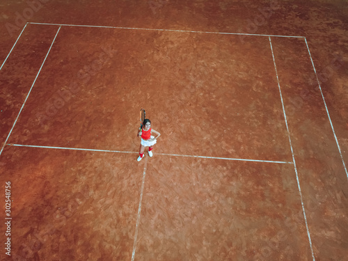 A young woman with a tennis racket in her hand stands in the center of a tennis court and looks into the camera. Top view, aerial view