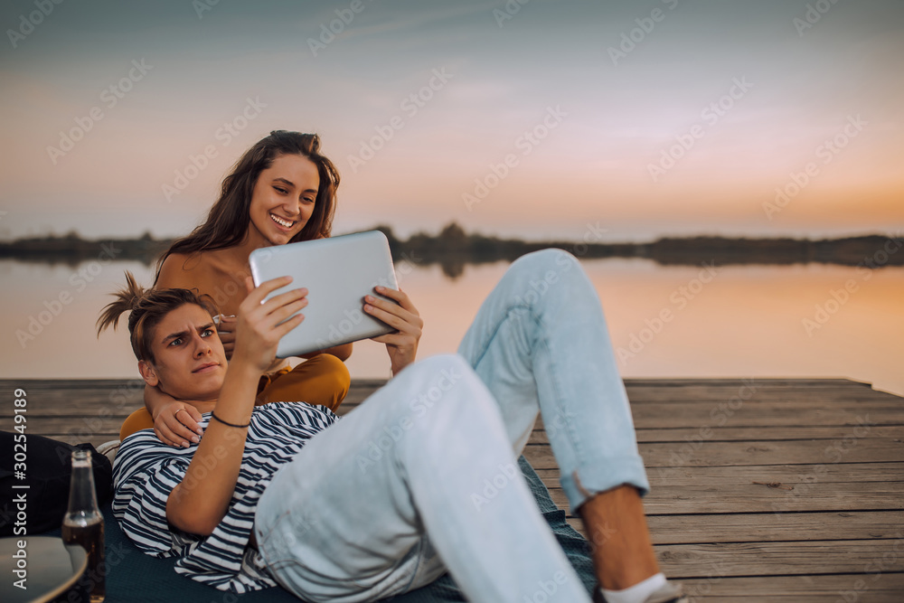 Young couple watching videos or sharing media content from a tablet, while sitting by the river Young couple watching videos or sharing media content from a tablet, while sitting by the river