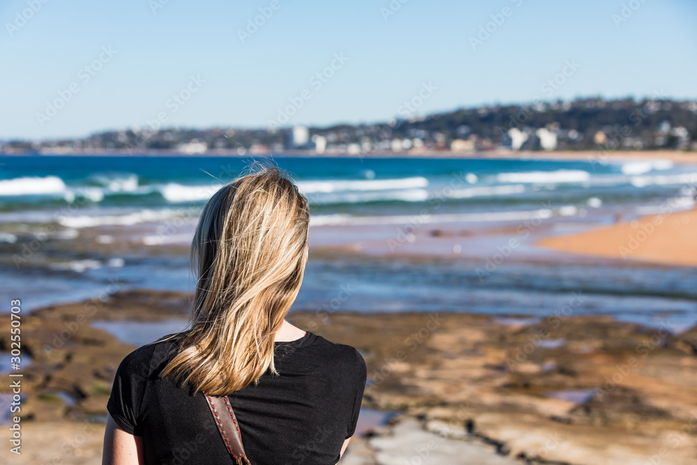 A blond woman standing and looking over the shore and headland at the beach on a blue sky sunny day.