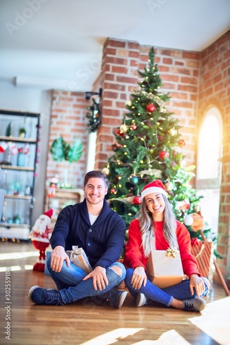 Young couple wearing santa claus hat sitting on the floor around christmas tree at home with a happy and cool smile on face. Lucky person.
