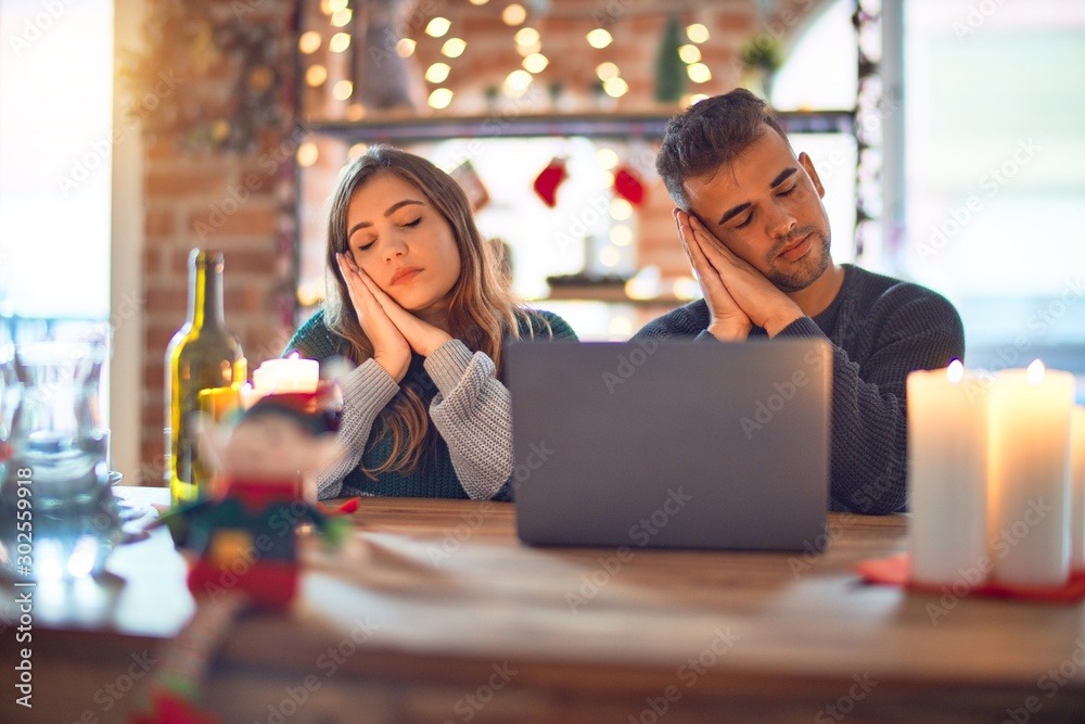 Young beautiful couple sitting using laptop around christmas decoration at home sleeping tired dreaming and posing with hands together while smiling with closed eyes.