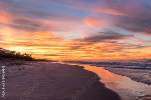 Spectacular orange sunrise seascape at Costa del Sol  El Salvador.   Horizontal orange clouds and reflection over the waves approaching the sand beach. Palm threes at the point where light rays origina