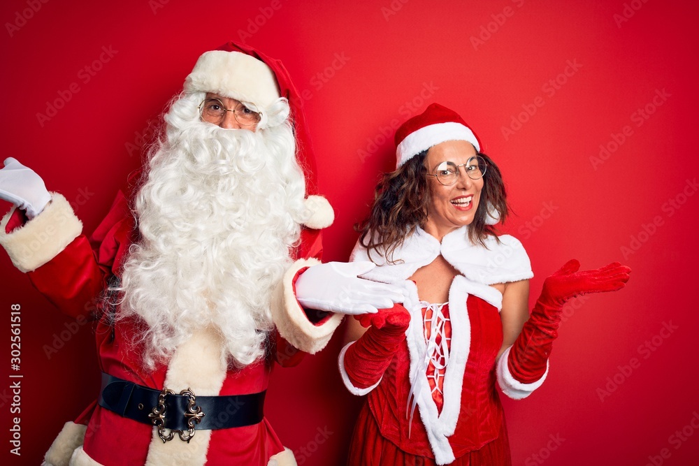 Middle age couple wearing Santa costume and glasses over isolated red background smiling showing both hands open palms, presenting and advertising comparison and balance Middle age couple wearing Santa costume and glasses over isolated red background smiling showing both hands open palms, presenting and advertising comparison and balance