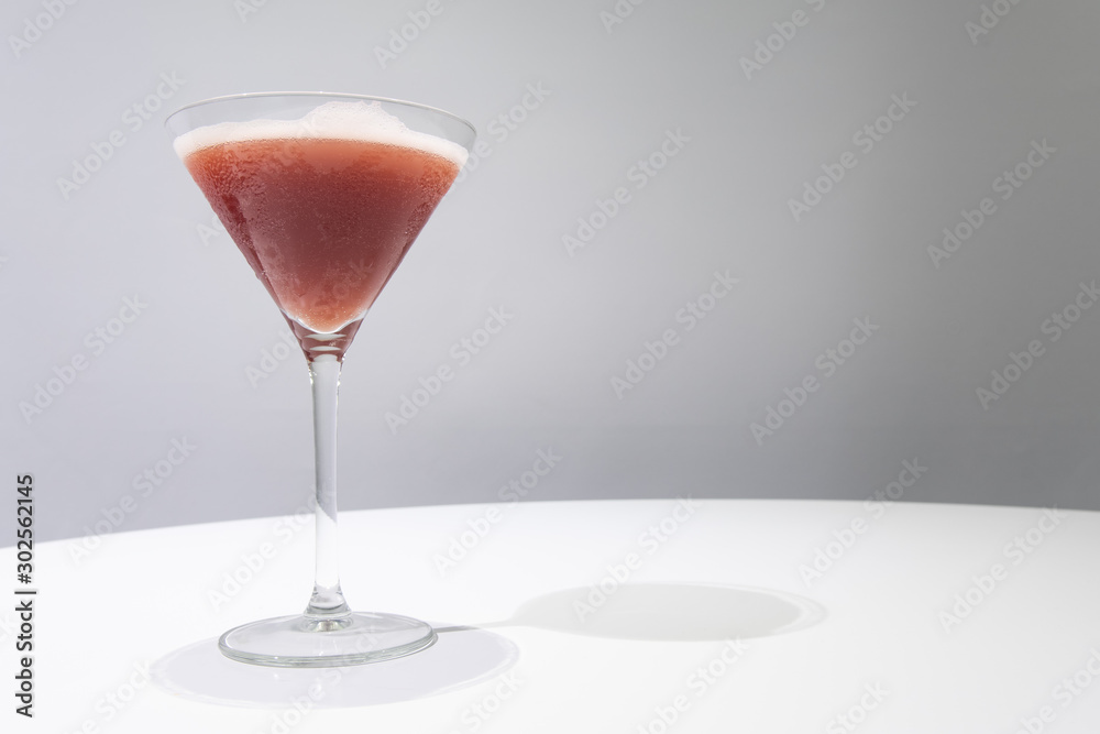 Cocktail glass with red colored cocktail in it on white table, gray background. Drink concept. Sangria cocktail. Selective focus. Copy space. Horizontal. Cocktail glass with red colored cocktail in it on white table, gray background. Drink concept. Sangria cocktail. Selective focus. Copy space. Horizontal.