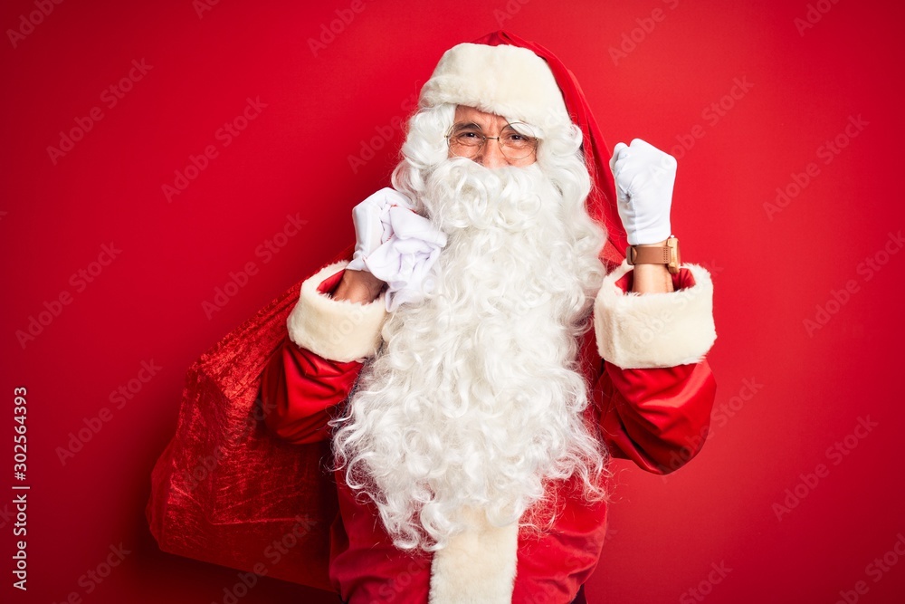 Middle age man wearing Santa costume holding sack with gifts over isolated red background screaming proud and celebrating victory and success very excited, cheering emotion