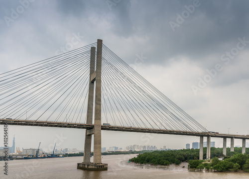 Ho Chi Minh City, Vietnam - March 12, 2019: Long Tau and song Sai Gon rivers meeting point. Landscape with H-shaped pylon of Phu My suspension bridge in center under gray cloudscape. Brown water. © Klodien