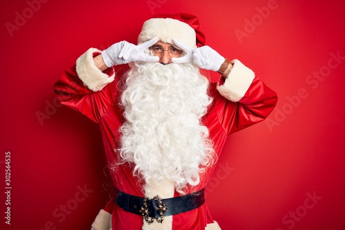 Middle age handsome man wearing Santa costume standing over isolated red background Doing peace symbol with fingers over face, smiling cheerful showing victory