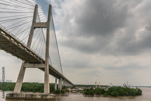 Ho Chi Minh City, Vietnam - March 12, 2019: Song Sai Gon river. One H-shaped pylon of Phu My suspension bridge and east side on ramp under gray cloudscape. Brown water. Long Tau river on right. © Klodien