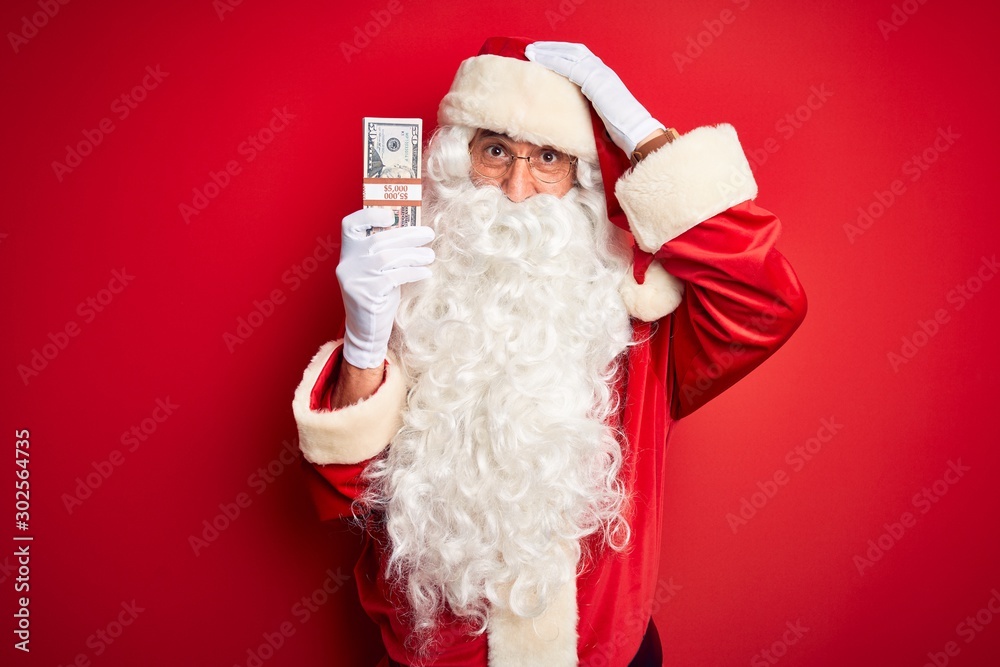 Middle age man wearing Santa Claus costume holding dollars over isolated red background stressed with hand on head, shocked with shame and surprise face, angry and frustrated. Fear and upset