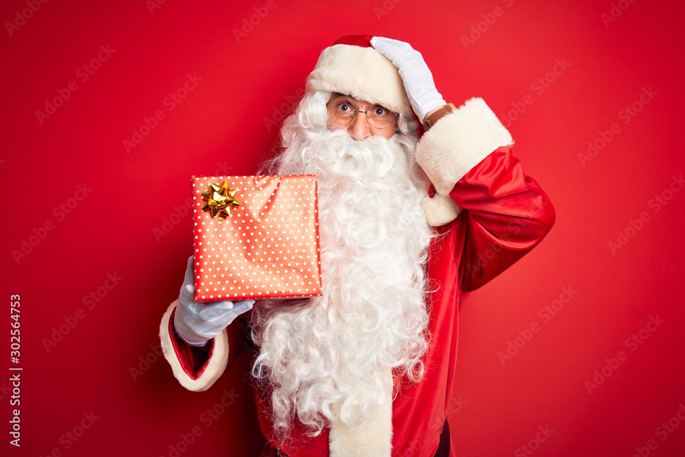 Middle age man wearing Santa Claus costume holding gift over isolated red background stressed with hand on head, shocked with shame and surprise face, angry and frustrated. Fear and upset for mistake.