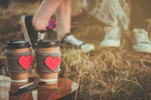 Fototapeta Naklejka Na Ścianę i Meble -  Romantic couple hugging while having picnic in sunset light. Coffee cup cozy knitted sleeve with felt red heart.
