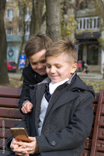 Two boys in business clothes on a bench in the park on an autumn day. Children watch something interesting on a smartphone. One smiles and points a finger at the screen, wanting to call attention