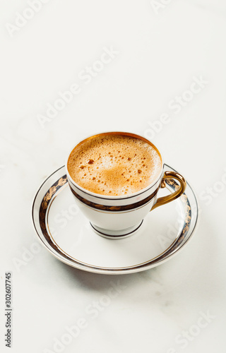 Cup of fresh americano or espresso coffee with golden foam froth on pile of brown raw coffee beans on white marble table background. Morning hot drink  coffee break  cope space