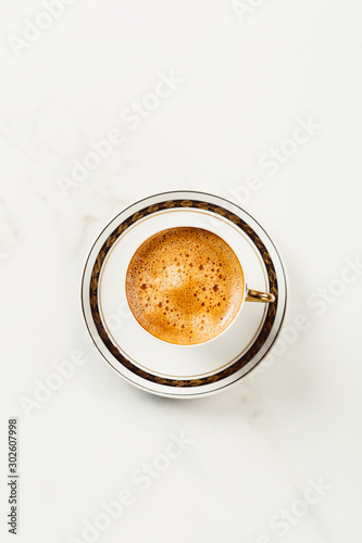 Cup of fresh americano or espresso coffee with golden foam froth on pile of brown raw coffee beans on white marble table background. Morning hot drink, coffee break, cope space