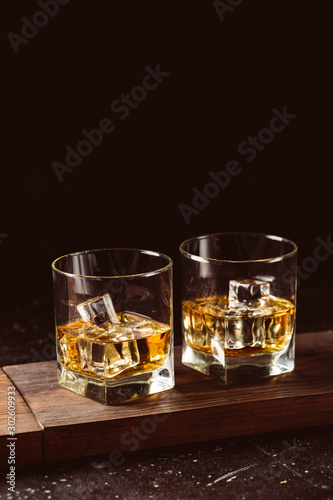 Two glasses of whiskey with ice cubes served on wooden board. Dark background, glossy golden color hard liquor on countertop with highlight. Night life bar relaxation concept