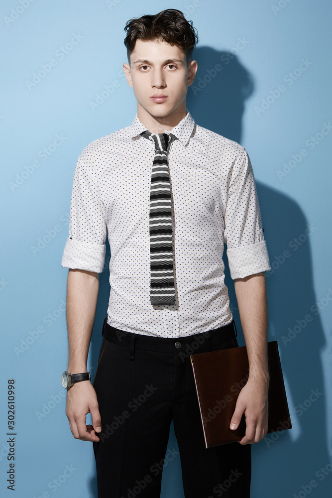 Fototapeta premium Medium full shot of a young dark haired man in a black trousers and white printed shirt with a knit tie with black, gray and white stripes. The man with a black watch is holding a leather folder. 