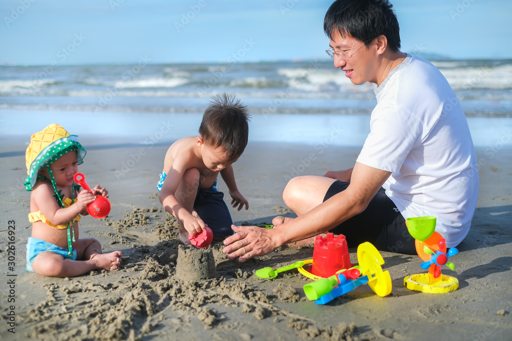 Fototapeta premium Asian father playing sand with baby girl daughter and toddler boy son, Dad playing beach toys with kids, Family travel, Summer beach vacation, Selective focus and father
