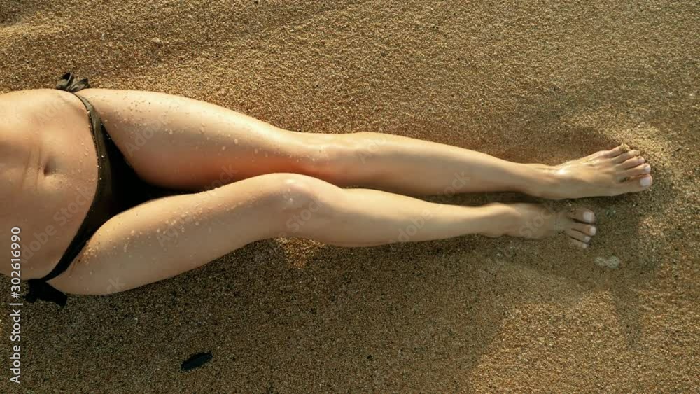 Close-up shot of legs of beautiful young woman sitting on the beach on a sunny day. Sea waves washing over tanned female feet. Spain. Slow motion. HD Close-up shot of legs of beautiful young woman sitting on the beach on a sunny day. Sea waves washing over tanned female feet. Spain. Slow motion. HD