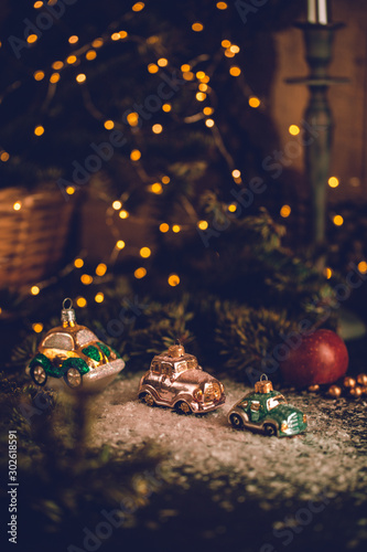 Christmas tree ornaments, tiny car on snowy table under the fir tree and bokeh lights with white candle in the background. Beautiful mysterious village decoration, greeting card, new year celebration 