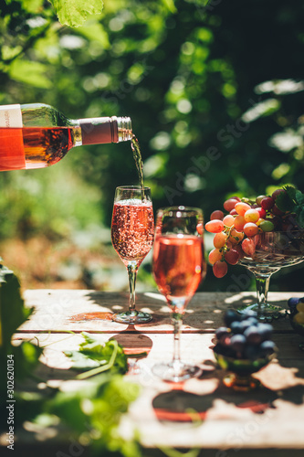 Rose wine tasting, glass of rose wine poured from bottle outdoors in garden party in vineyard, ripe grapes on wooden table, sunlight, harvest time, copy space