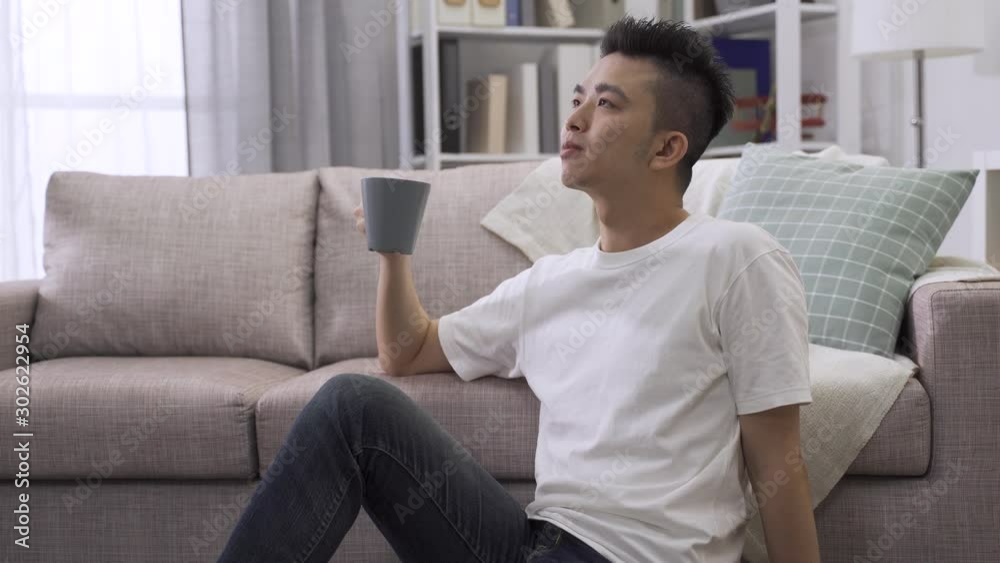 Handsome young asian chinese man sitting on floor relaxed drinking cup of coffee and smiling at home living room. confident male relax leaning on couch with water. healthy lifestyle concept. Handsome young asian chinese man sitting on floor relaxed drinking cup of coffee and smiling at home living room. confident male relax leaning on couch with water. healthy lifestyle concept.