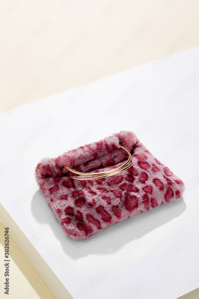 Object shot of a pink leopard print faux fur bag with metallic semicircular bag handles, lying on a white surface. The bag is casting a shadow.