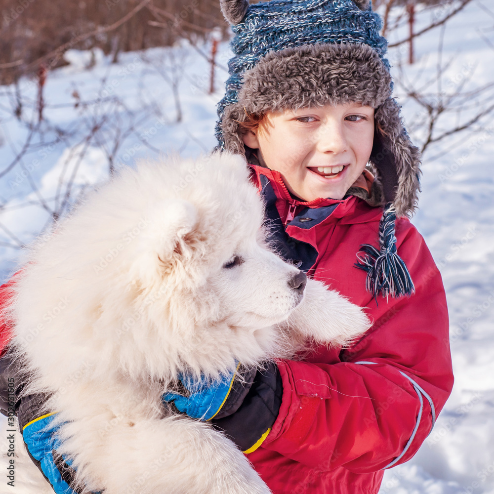 A boy in a hat with ears is hugging a samoyed puppy outdoors in the winter. Two cute children - friendship of man and dog A boy in a hat with ears is hugging a samoyed puppy outdoors in the winter. Two cute children - friendship of man and dog
