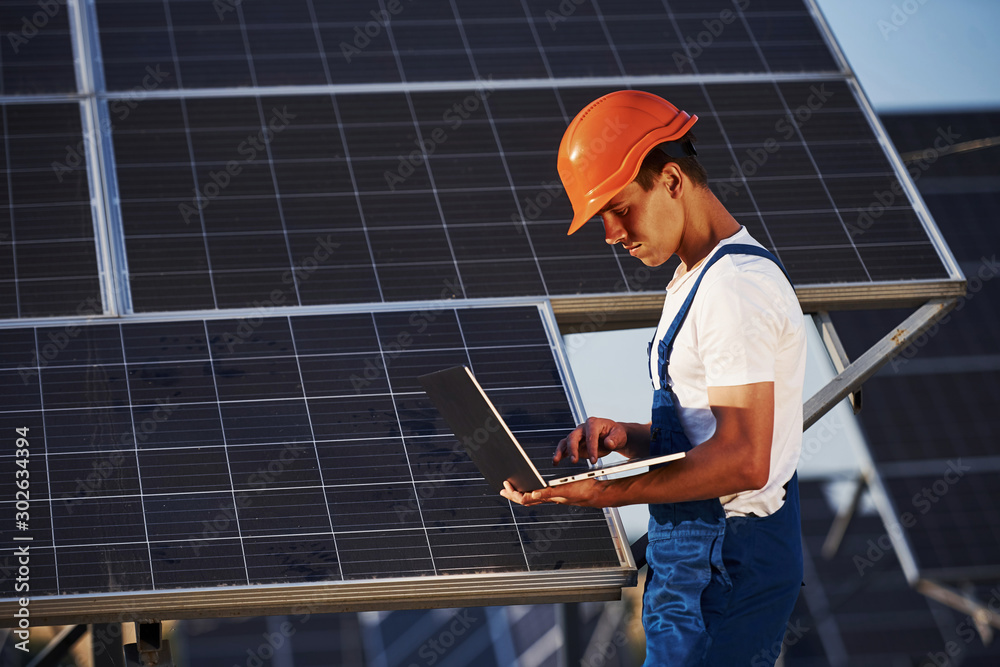 Using laptop. Male worker in blue uniform outdoors with solar batteries at sunny day Using laptop. Male worker in blue uniform outdoors with solar batteries at sunny day