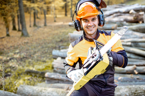 Portrait of a cheerful professional lumberjack in protective workwear standing with a chainsaw on a pile of logs in the forest