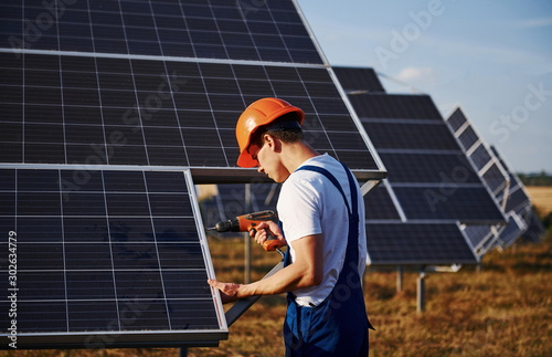 Using cordless screwdriver. Male worker in blue uniform outdoors with solar batteries at sunny day
