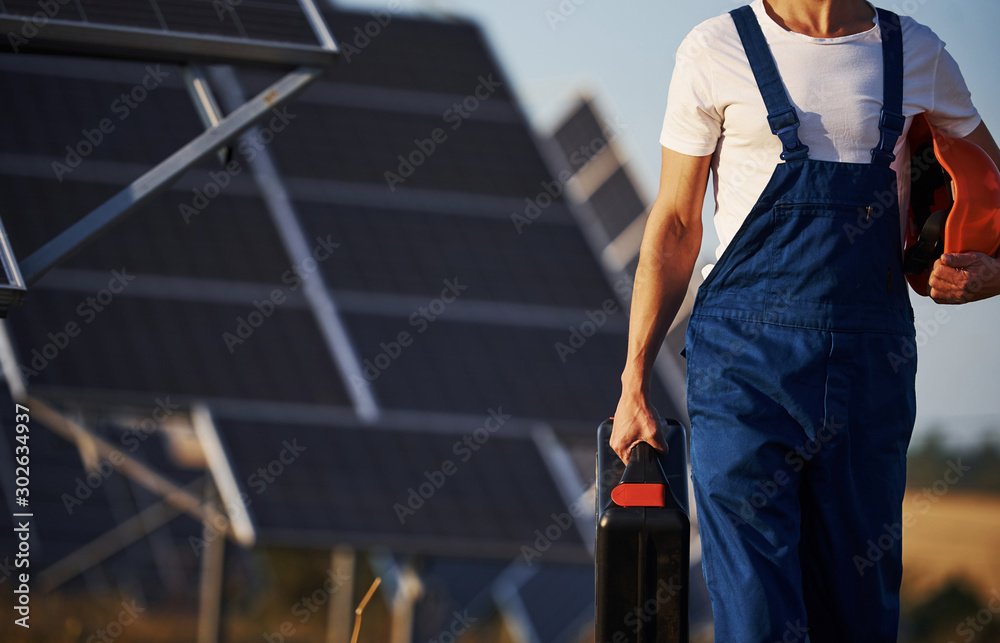 Holds case with equipment in hand. Male worker in blue uniform outdoors with solar batteries at sunny day