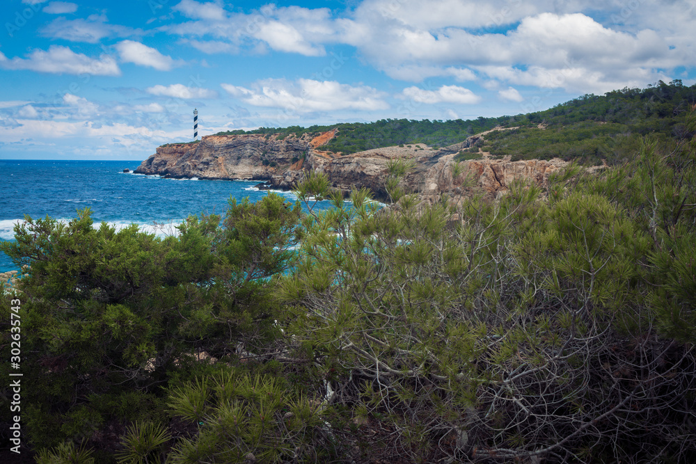 Moscarter lighthouse on the Ibiza island in the Mediterranean sea on top of a cliff shot from coast, wide landscape with rocky coast in foreground