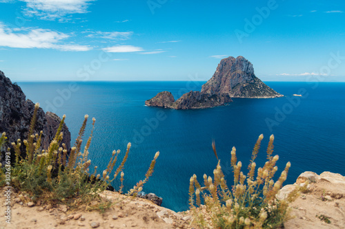 Fototapeta Naklejka Na Ścianę i Meble -  View of famous mysterious island Es Vedrà from famous view point on cliffs above beach Cala D'Hort beautiful island of Ibiza, Spain, scenic blue sea, horizon sky, plants and cliff rocks in foreground