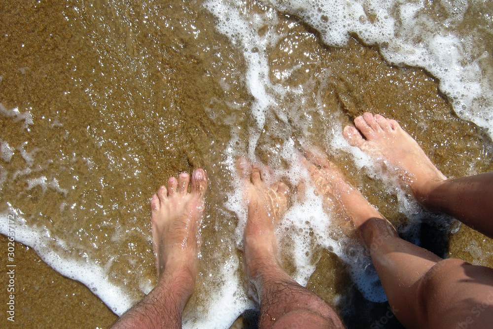 Fototapeta premium The legs of a man and a woman stand on the sandy shore of the sea and they are slightly doused with water
