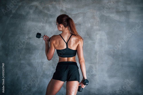 Young fitness woman is in the gym near wall with dumbbells in hands